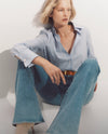 A woman with blonde hair sits on the floor, wearing a striped button-up shirt and flared jeans, with a contemplative expression on her face.