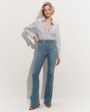 A woman with blonde hair stands in a plain white background, wearing a light blue button-up shirt and high-waisted flared jeans.