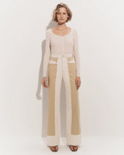 A young woman with long blonde hair stands in a plain white background, wearing a beige sweater and wide-leg tan pants.