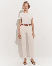A young woman with blonde hair stands in a plain white background, wearing a white t-shirt, beige pants, and sandals.