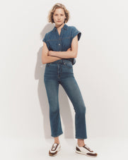 A young woman with blonde hair stands against a plain white background, wearing a denim shirt and high-waisted jeans, with her arms crossed and a neutral expression on her face.