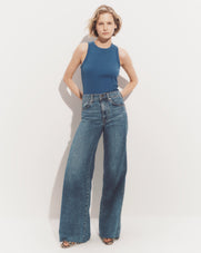 A young woman with blonde, wavy hair stands against a plain white background, wearing a blue sleeveless top and wide-leg denim jeans.
