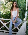 A young woman with long dark hair sits on a wooden bench surrounded by lush tropical foliage in the background.