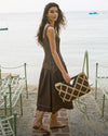 A young woman with long, curly hair stands on a beach, holding a patterned bag, with a body of water and boats visible in the background.