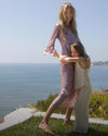 A woman in a shimmering pink dress hugs a young girl in a white top and pants, with a vast blue ocean and distant coastline in the background.