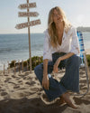 A woman in a white top and blue jeans sits on a beach chair in the foreground, with a signpost pointing to "Malibu," "Summering," and "Veronica Beard" behind her, and the ocean and sky in the background.
