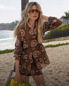 A blonde woman in a patterned brown outfit stands on a sandy beach with the ocean and palm trees in the background.