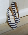 Striped ballet flats with ankle straps rest on a wooden floor in front of a white wall.