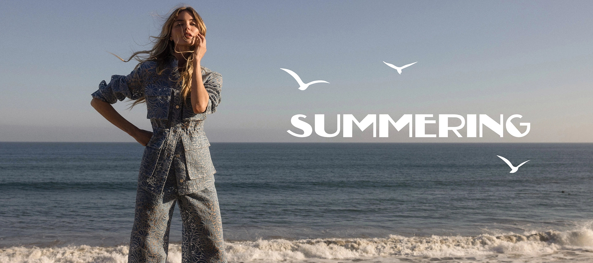 A woman in a denim outfit stands on a beach with the ocean and sky in the background, with the word "SUMMERING" and three seagulls superimposed.