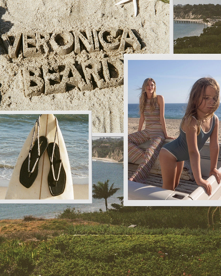 The foreground features a sandy beach with "VERONICA BEARD" carved into it, a surfboard with flip-flops, and lush green grass, while the background shows two young women on a beach with the ocean and cliffs.