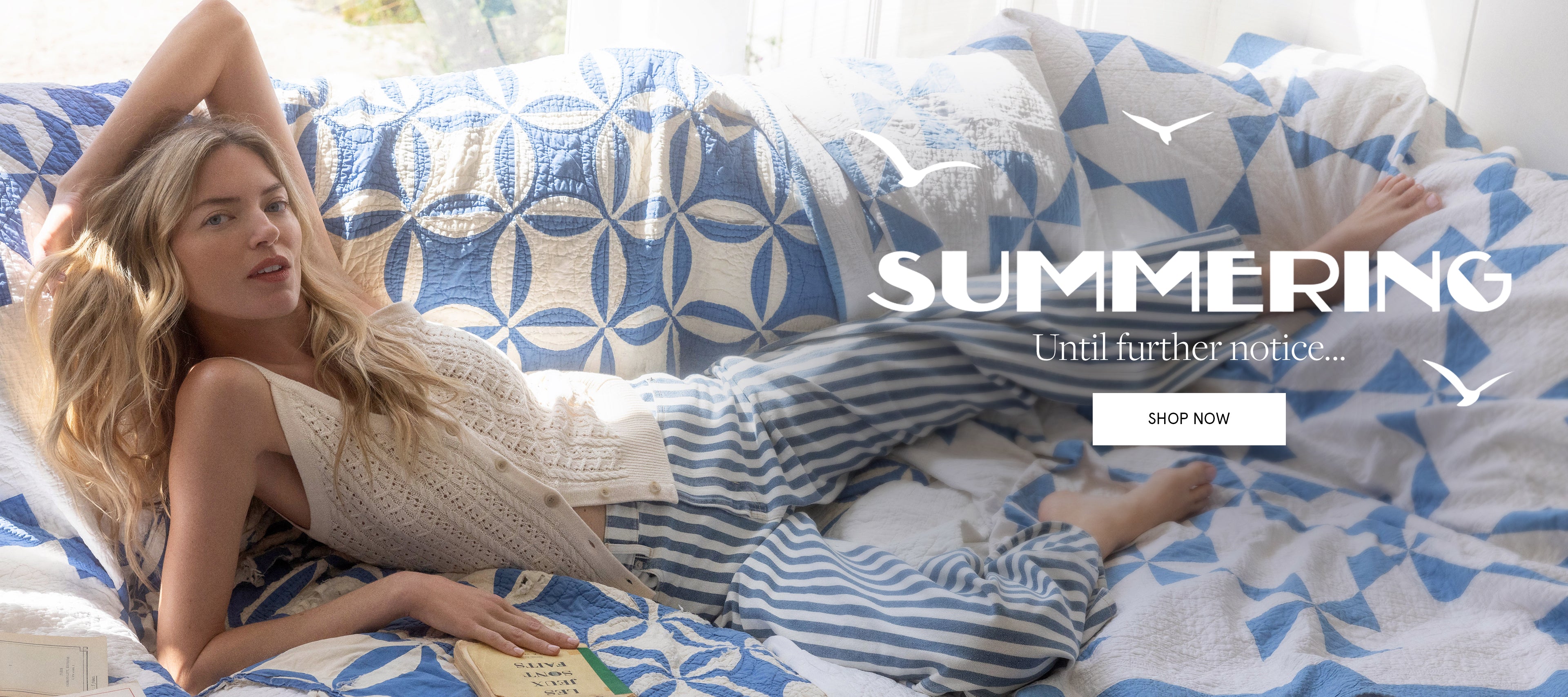 A blonde woman in striped pajamas reclines on a bed covered in blue and white patterned quilts, with the word "SUMMERING" and a "SHOP NOW" button overlaid on the background.