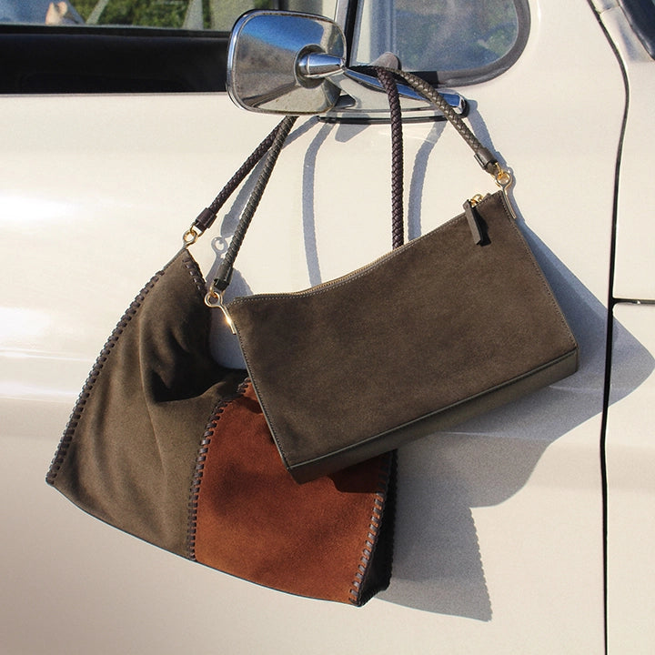 Two suede handbags, one larger and patchwork, hang from a car door handle against a blurred background of greenery and sky.