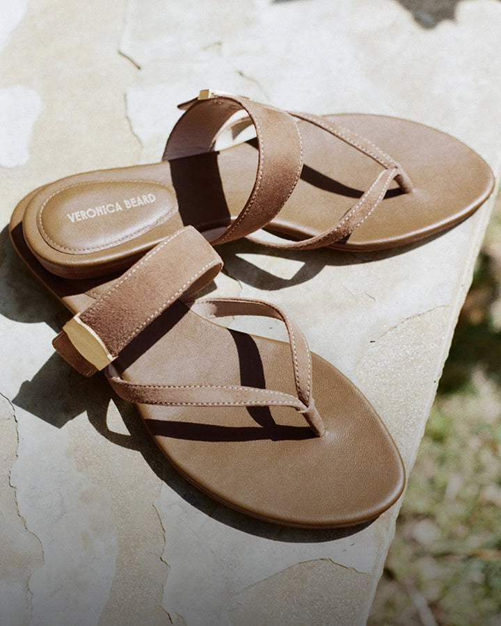 A pair of brown leather thong sandals with gold buckles rests on a textured stone surface, with a blurred background of greenery.