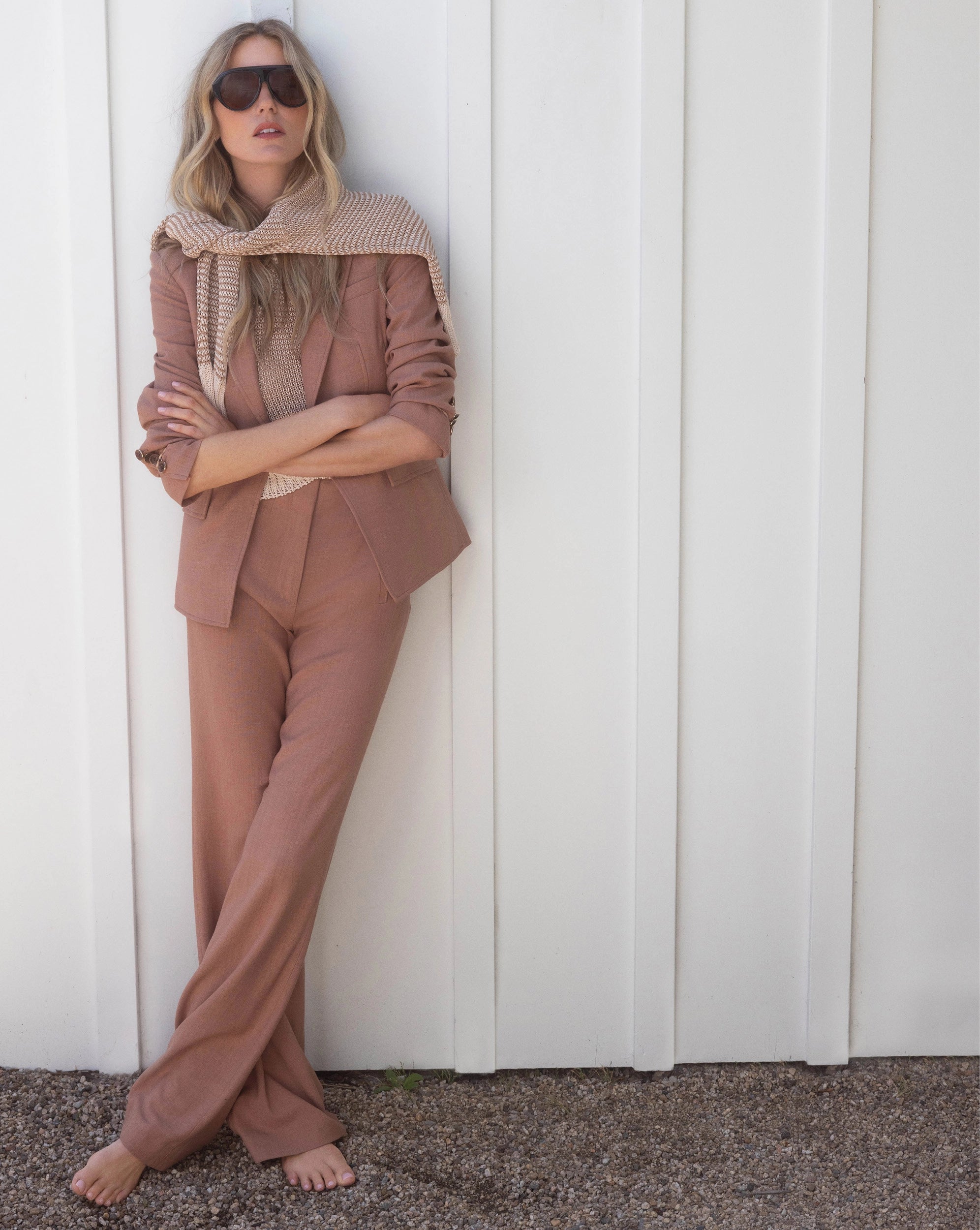 A woman in a tan suit and scarf leans against a white paneled wall, with gravel in the foreground.