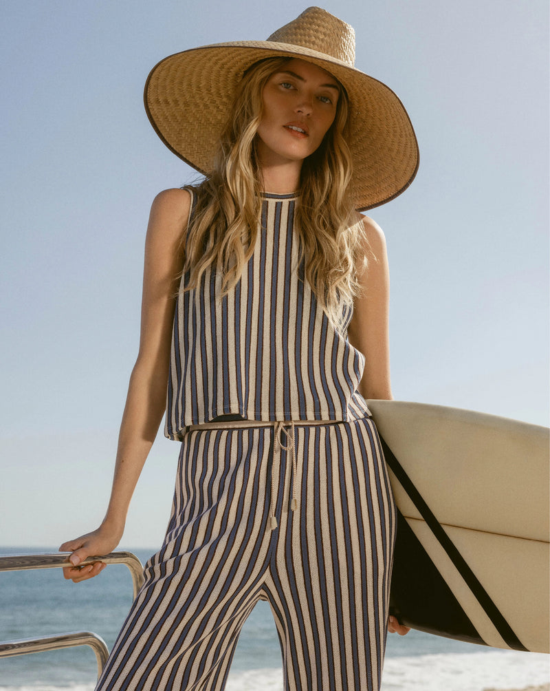 A woman in a striped outfit and wide-brimmed hat holds a surfboard against a backdrop of the ocean and clear sky.