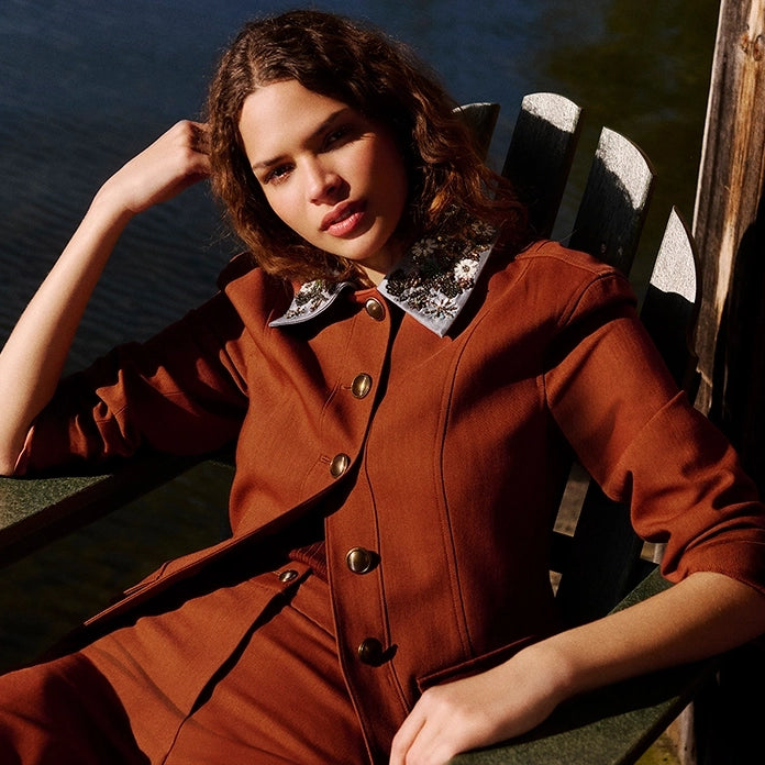 A young woman in a brown suit sits in a chair by the water, with a wooden dock to her right.