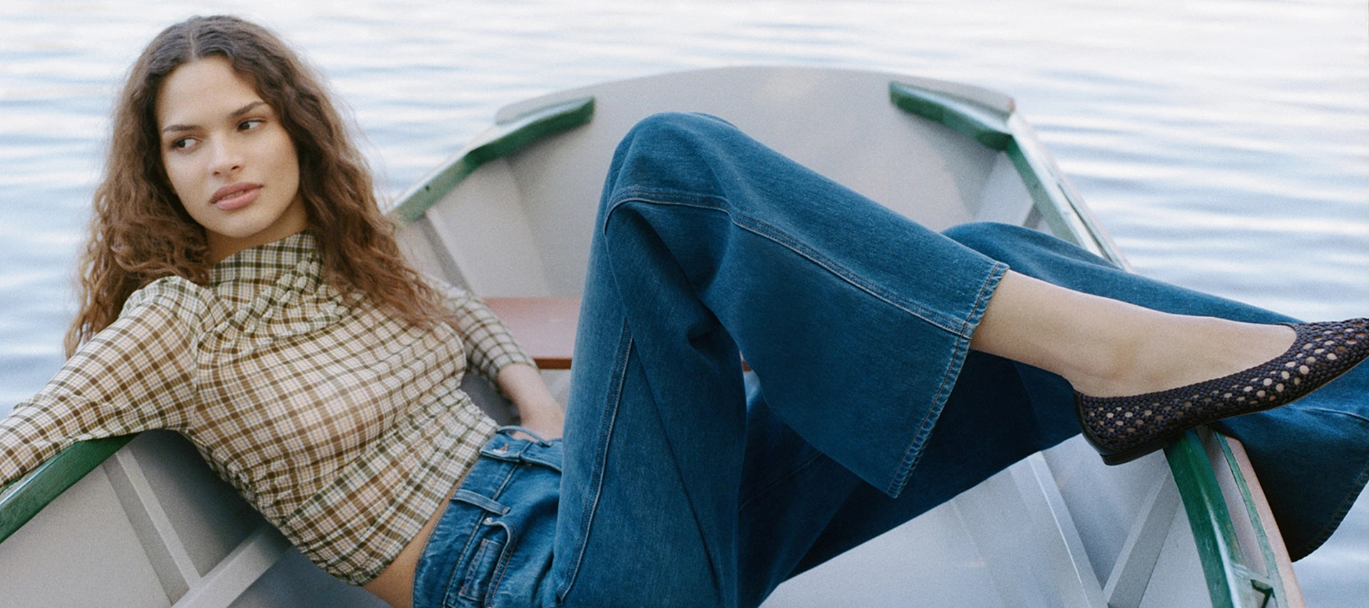 A woman in a checkered top and blue jeans relaxes in a boat on calm water.