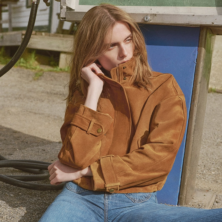 A young woman in a brown jacket and blue jeans sits on the ground in front of a blue structure, with a hose and gravel in the foreground and a building in the background.