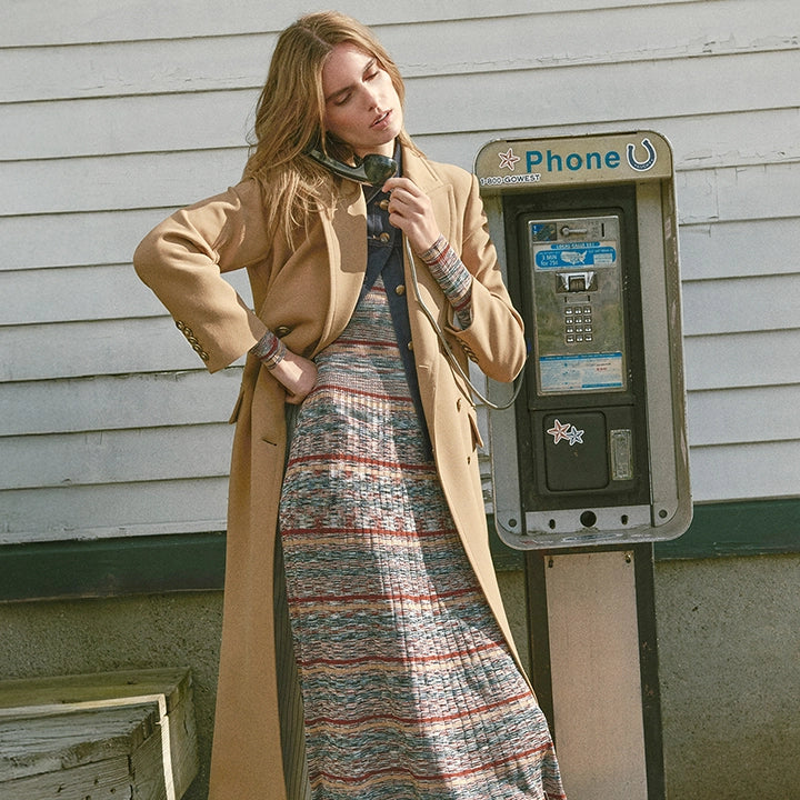 A woman in a long coat and patterned dress stands by a payphone in front of a weathered white building.