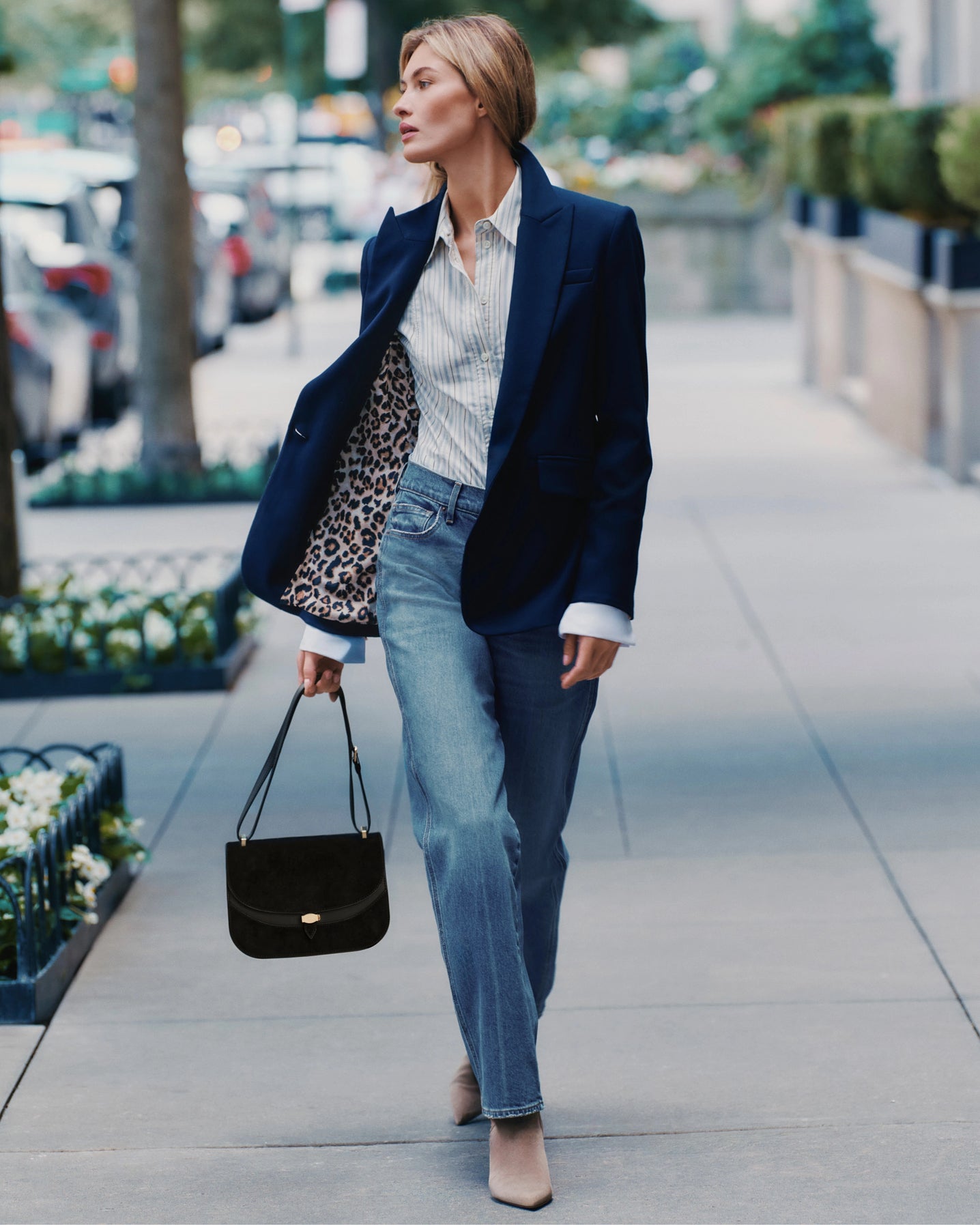 A woman in a navy blazer and jeans walks down a sidewalk with a black handbag, while cars and buildings are visible in the blurred background.