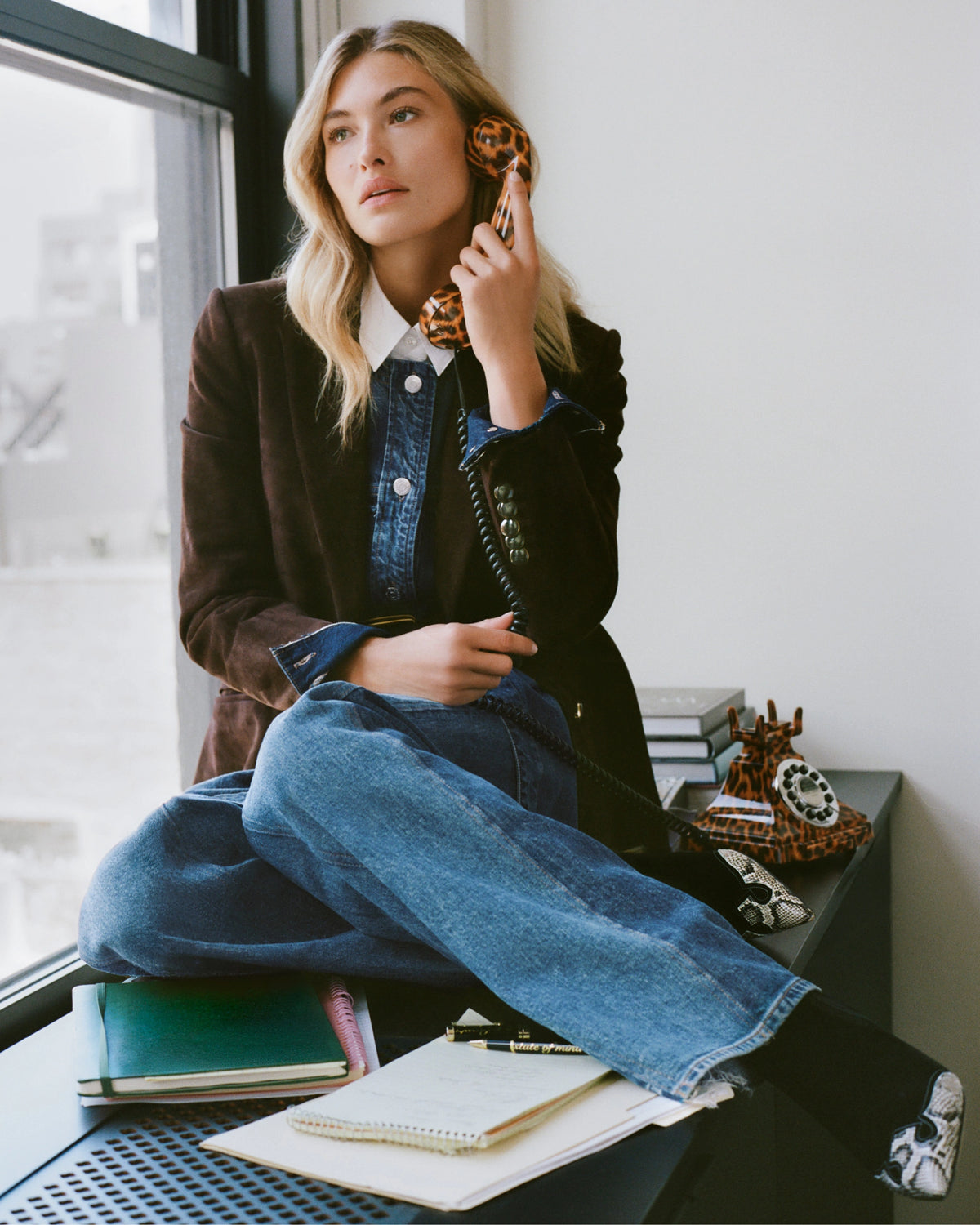 A blonde woman in a blazer and jeans sits on a desk holding a leopard-print phone, with a window and a stack of books in the background.