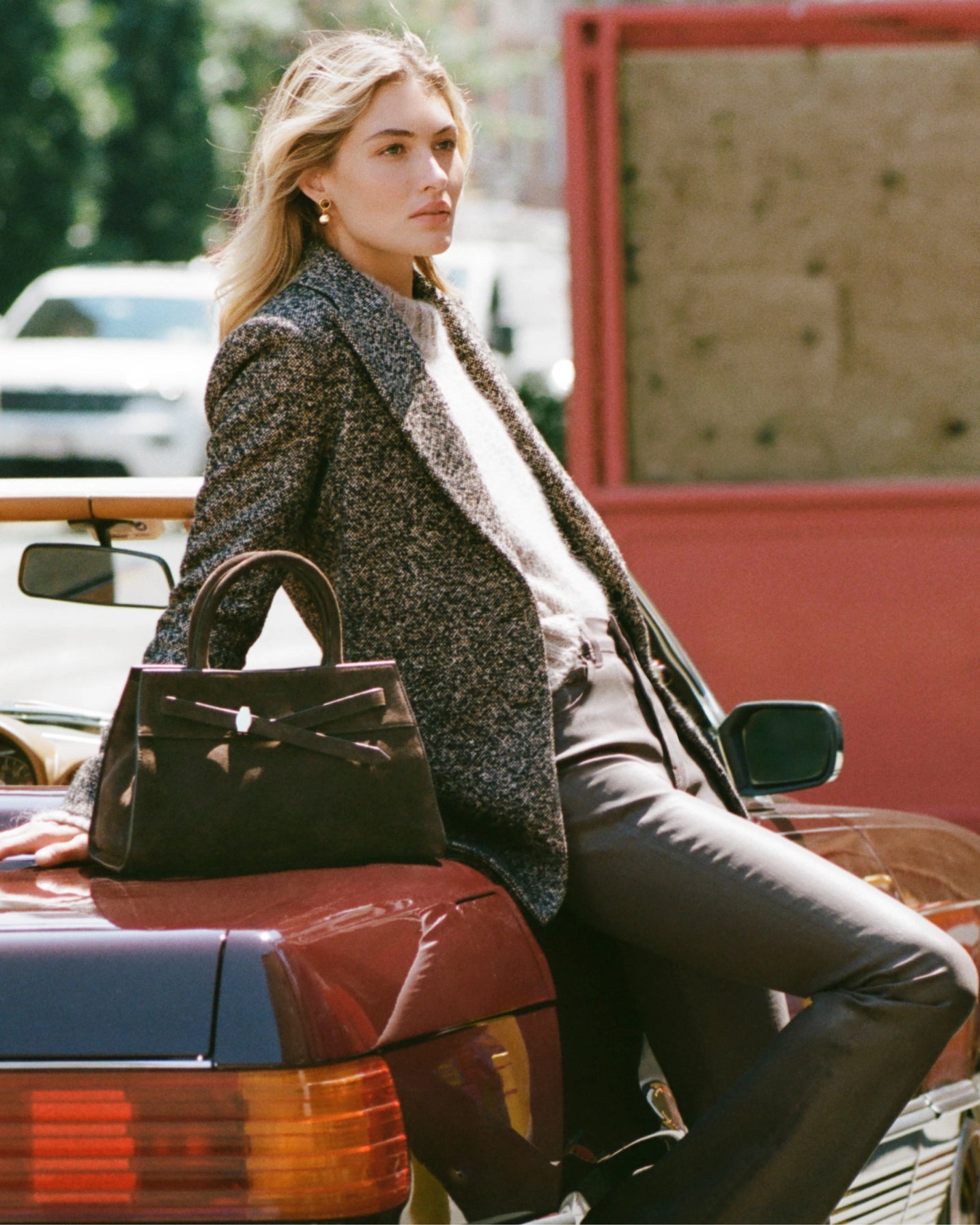 A woman in a tweed jacket and dark pants leans against a car with a brown handbag on its trunk, while a white car is visible in the blurred background.