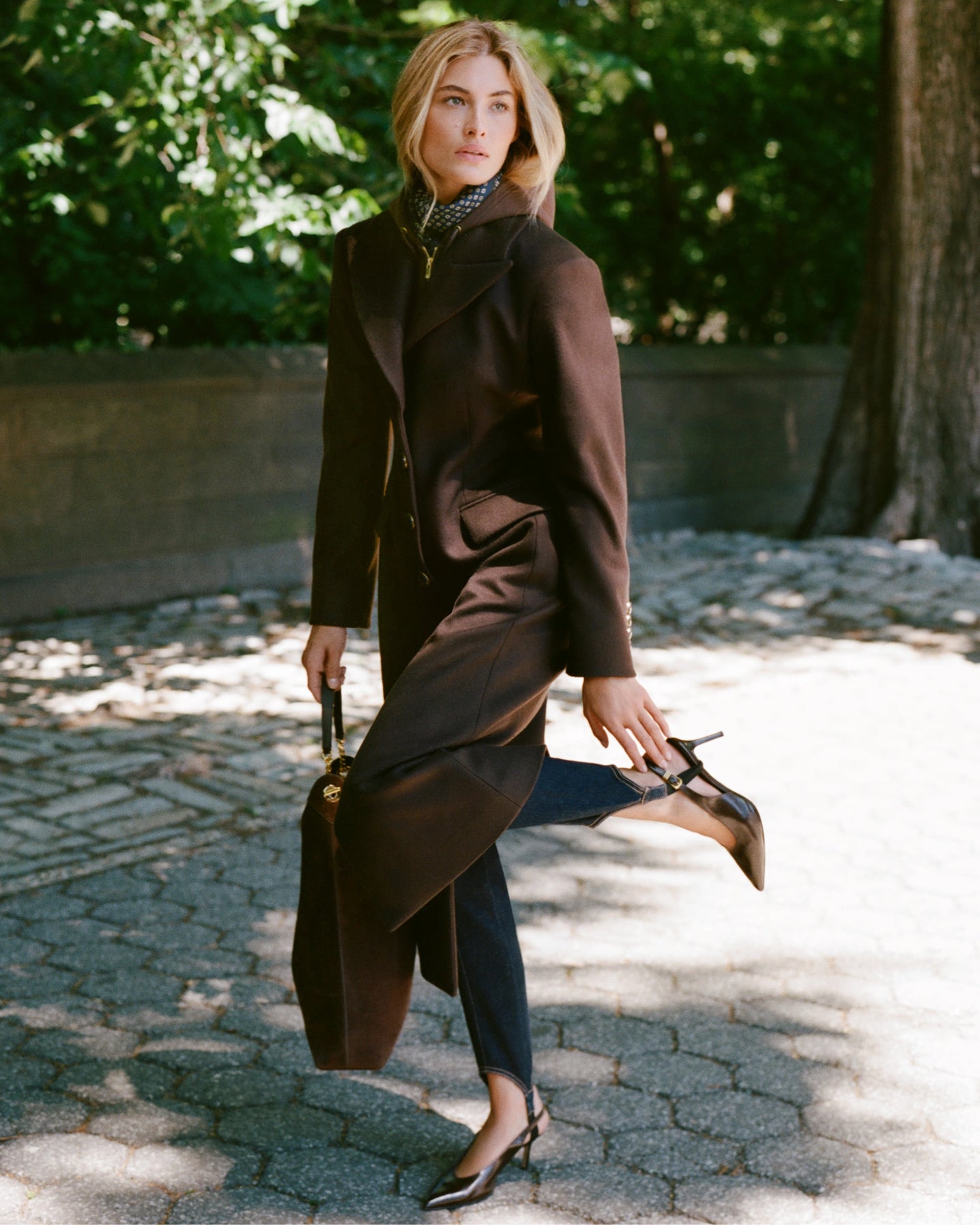 A woman in a brown coat and heels poses on a paved path with lush green trees in the background.