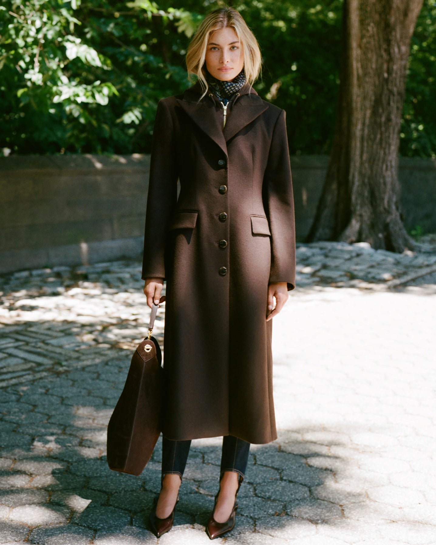 A woman in a long brown coat stands on a cobblestone path with dappled sunlight, while lush green trees and a stone wall form the background.