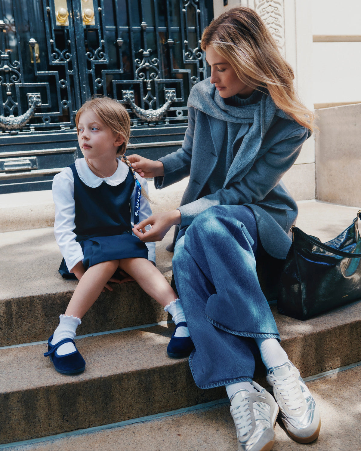 A young girl in a school uniform sits on steps in the foreground while a woman in a grey coat and jeans adjusts her hair, with a dark ornate gate in the background.