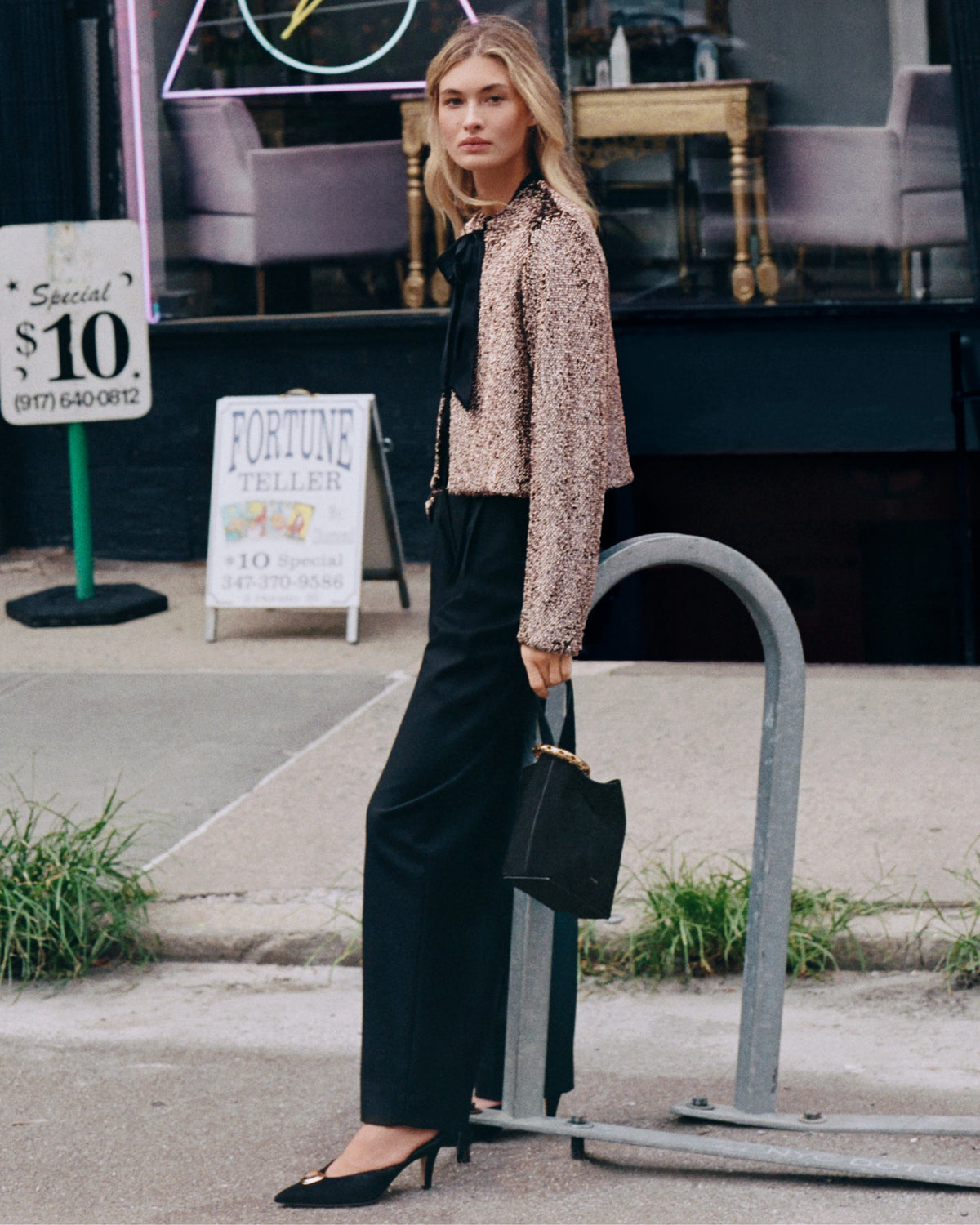 A woman in a sequined jacket and black pants stands on a sidewalk in front of a fortune teller's shop.