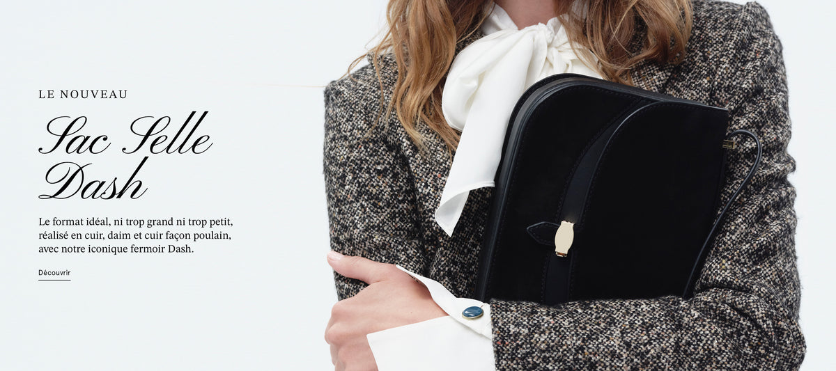 A person wearing a textured grey blazer and a white bow tie holds a black leather handbag against a plain white background.