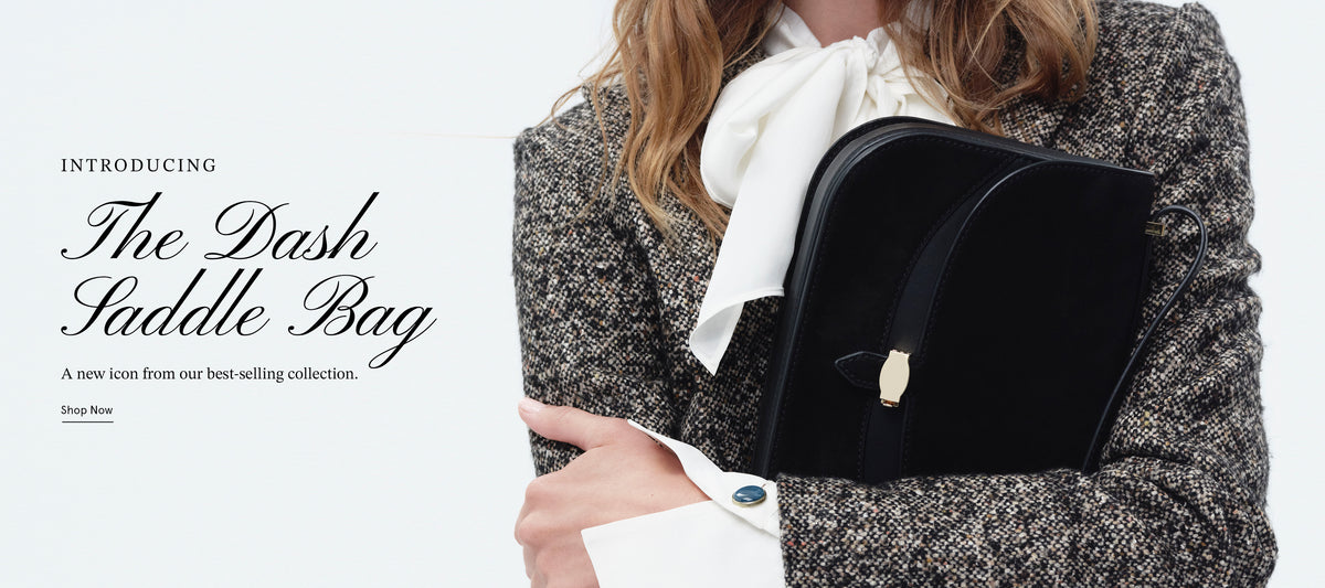 A person wearing a tweed blazer and white bow tie holds a black saddle bag against a plain white background.
