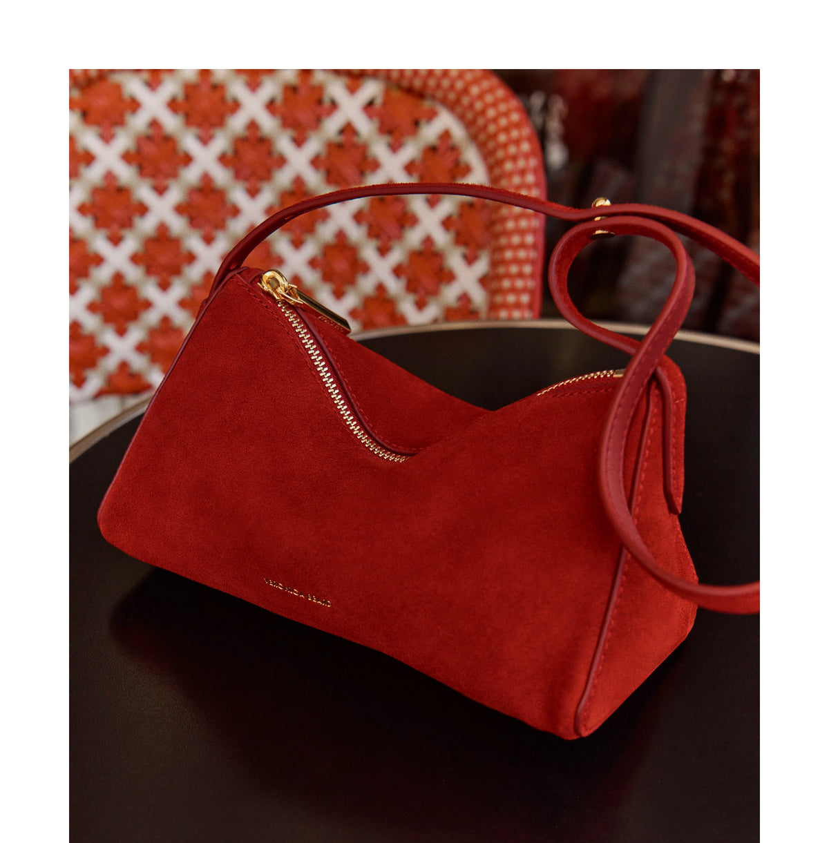 A red suede handbag sits on a dark table against a background of a red and white patterned surface.