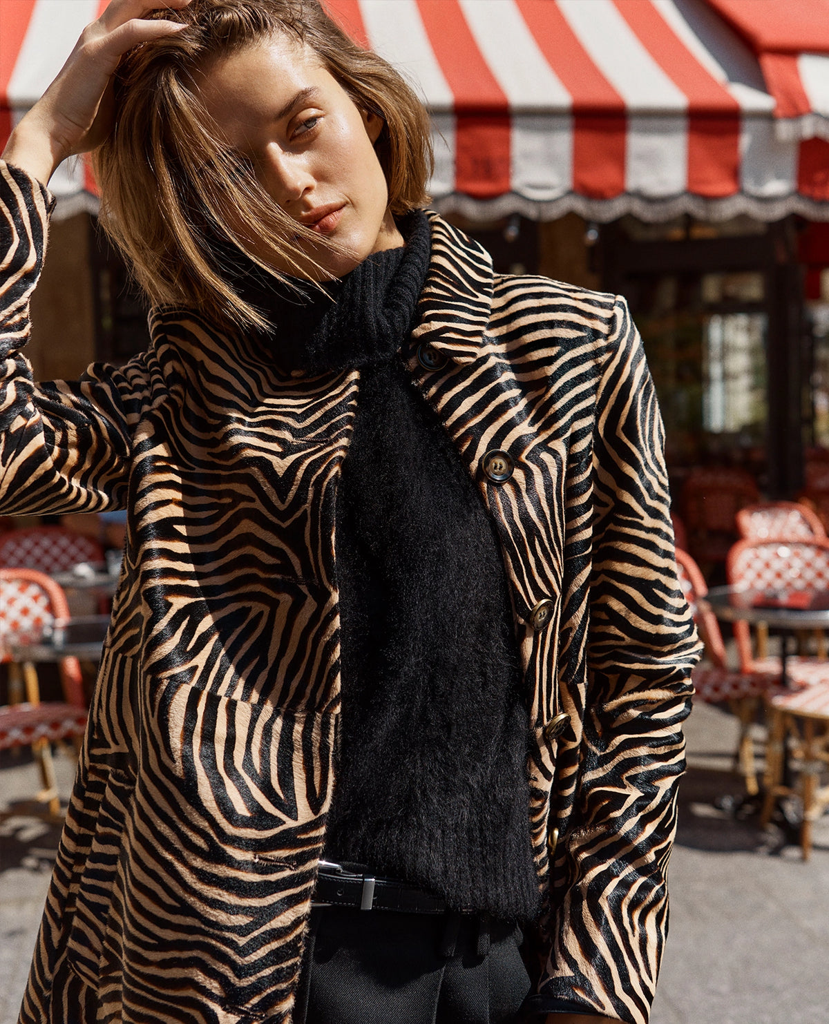 A woman wearing a zebra-print blazer and black turtleneck stands in the foreground against a background of a cafe with a red and white striped awning.