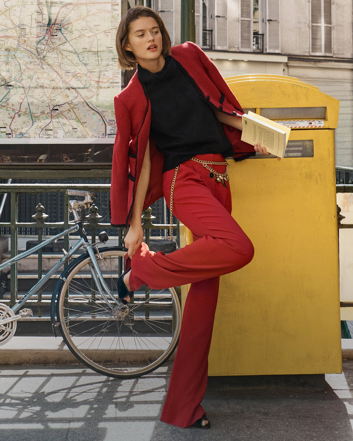 A woman in a red suit and black top leans against a yellow post box next to a bicycle, with a city map and buildings in the background.