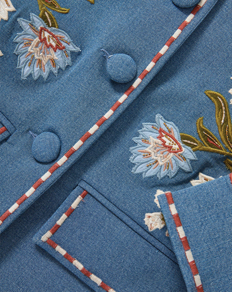 The image depicts a close-up view of a blue fabric or textile material with intricate floral embroidery patterns in various colors, including white, red, and gold. The background appears to be a solid blue color, while the foreground showcases the detailed embroidered designs.
