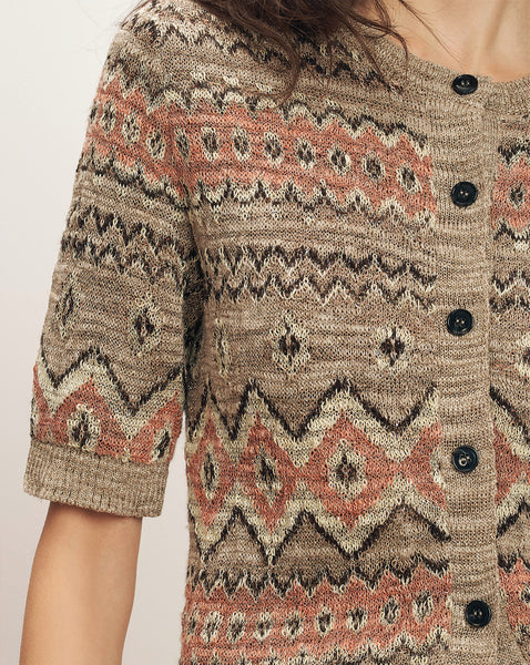 A close-up view of a person's upper body, showcasing a patterned knitted sweater with various geometric designs in shades of beige, brown, and coral.