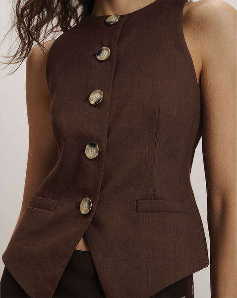 A close-up view of a person wearing a brown sleeveless jacket with large silver buttons, against a plain background.