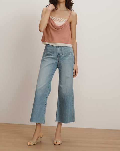 A young woman wearing a peach-colored tank top and light-wash denim culottes stands in a minimalist setting with a plain background.