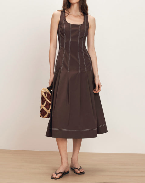 A woman wearing a brown sleeveless dress stands in a plain room, holding a patterned handbag.