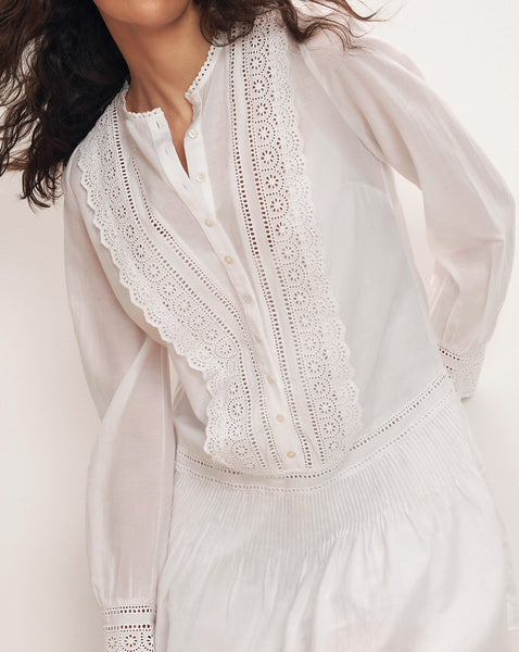 A close-up view of a woman wearing a white, lace-trimmed blouse with billowing sleeves against a plain, light-colored background.