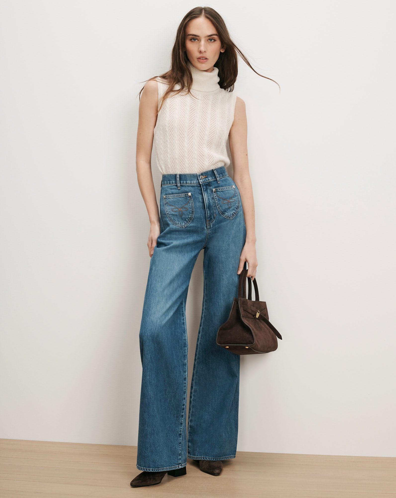 Veronica Beard Cream knit turtleneck, blue wide-leg jeans, brown suede handbag.