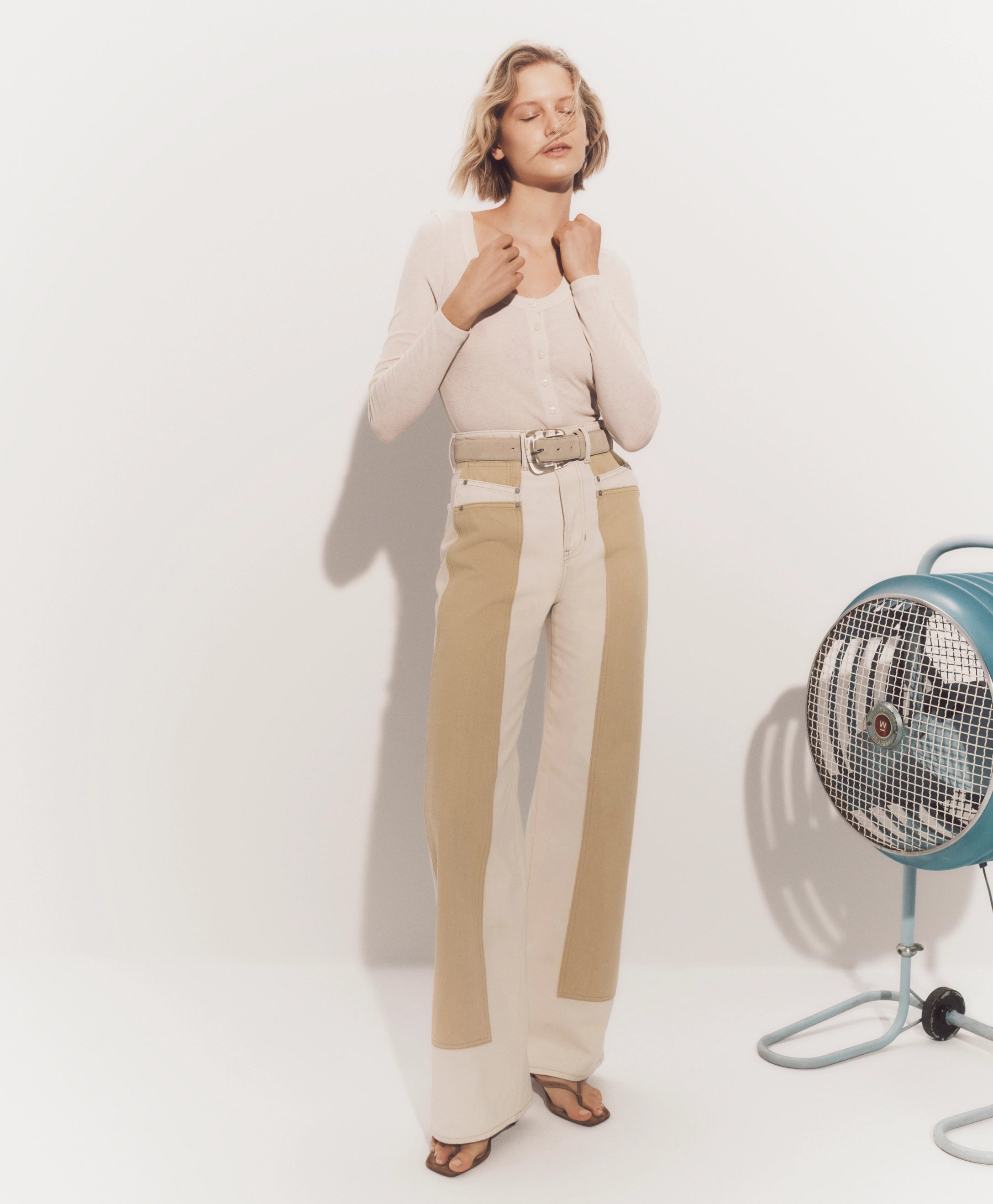 A woman with blonde hair stands in a neutral-colored room, wearing a beige sweater and pants, and holding her arms in front of her chest.