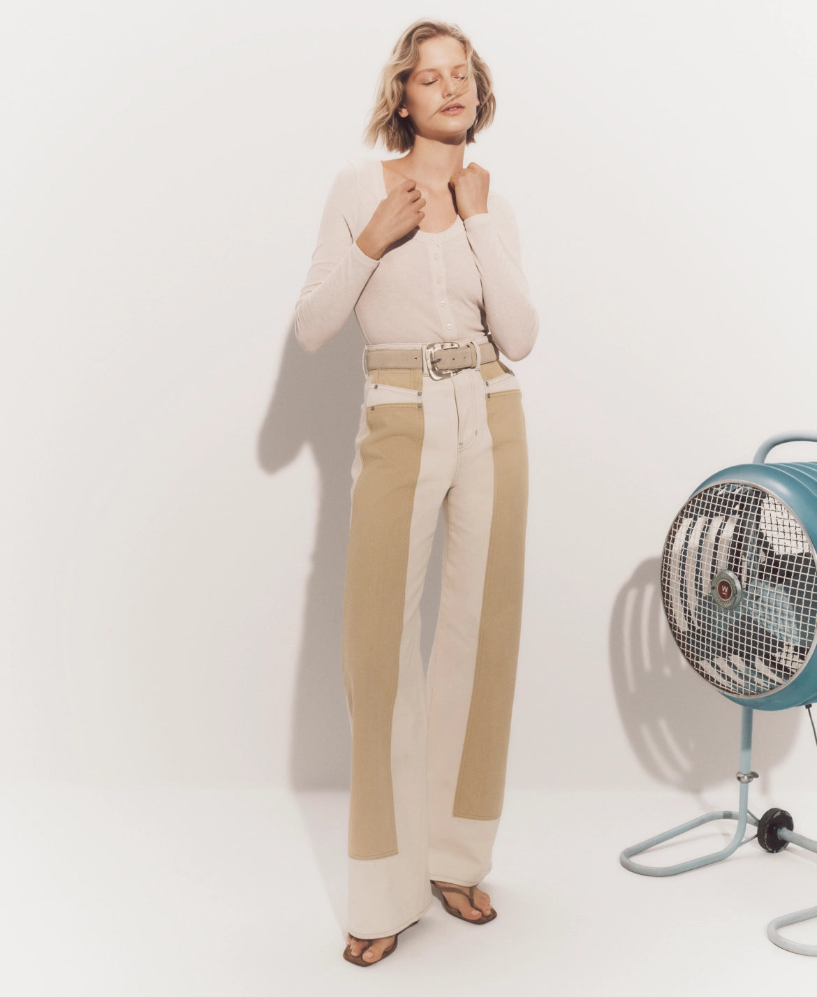 A woman with curly hair stands in a neutral-colored outfit, with a fan and disco ball visible in the background.